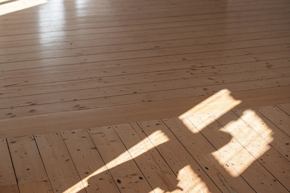 Empty room with bare sanded floorboards and window light, cleared of furniture during a hardwood floor refinishing job